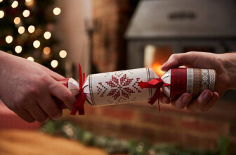 Two people pulling a decorated Christmas cracker in a cosy room with soft festive lights and a fireplace in the background.