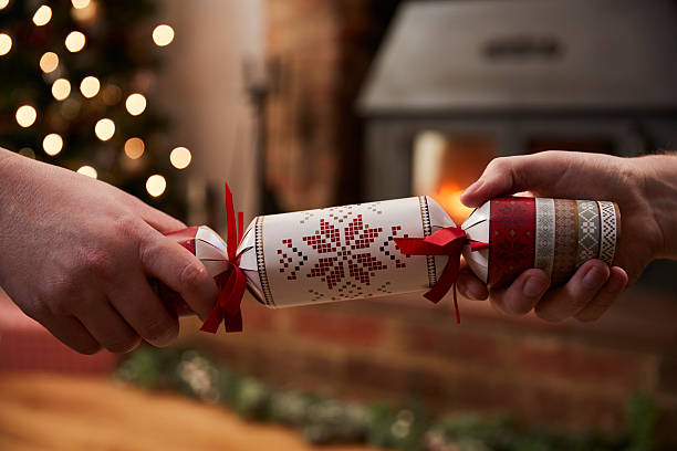 Two people pulling a decorated Christmas cracker in a cosy room with soft festive lights and a fireplace in the background.