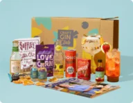 A Craft Gin Club subscription box with gin, mixers, snacks, a branded gin glass and a magazine arranged in front of the box on a light blue background.