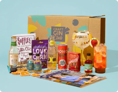 A Craft Gin Club subscription box with gin, mixers, snacks, a branded gin glass and a magazine arranged in front of the box on a light blue background.