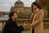 Man proposing to a woman on a stone bridge, holding an engagement ring in a city setting