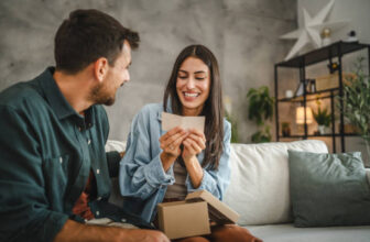 A woman sitting on a sofa opening a small gift box while smiling at the man beside her in a modern, clutter free living room