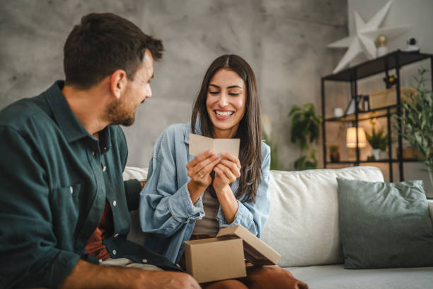 A woman sitting on a sofa opening a small gift box while smiling at the man beside her in a modern, clutter free living room