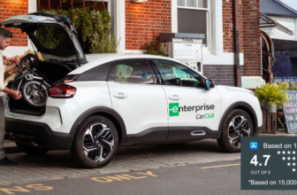 Man loading a folded wheelchair into the boot of a white Enterprise Car Club car parked outside a shop on a high street.