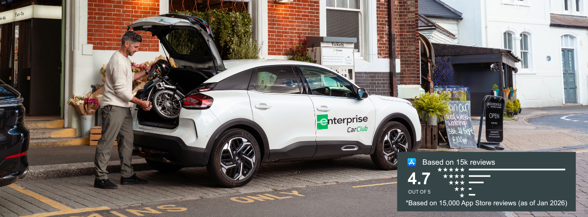 Man loading a folded wheelchair into the boot of a white Enterprise Car Club car parked outside a shop on a high street.